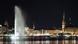 The large fountain spurts water with the city of hamburg illuminated in the background at night.